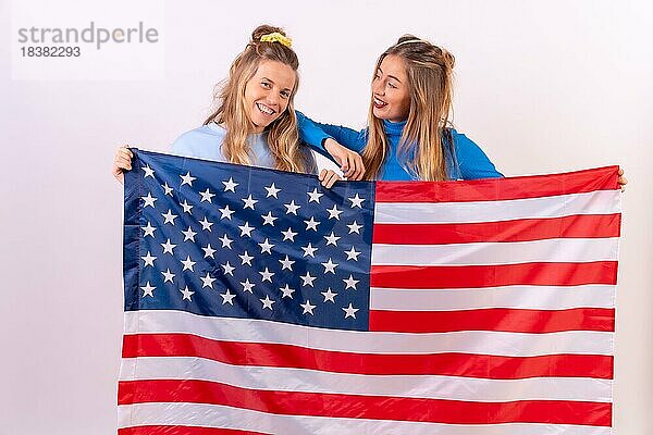 Zwei junge blonde kaukasische Frauen mit amerikanischer Flagge  lächelnd und Spaß habend  vor einem weißen Hintergrund