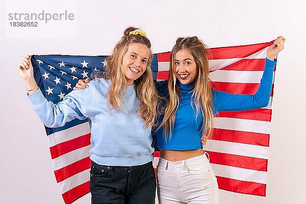 Zwei junge blonde kaukasische Frauen mit amerikanischer Flagge  lächelnd und Spaß habend  vor einem weißen Hintergrund