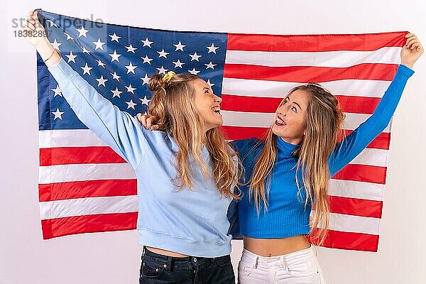 Zwei junge blonde kaukasische Frauen mit amerikanischer Flagge  lächelnd  während sie die Flagge halten  vor einem weißen Hintergrund