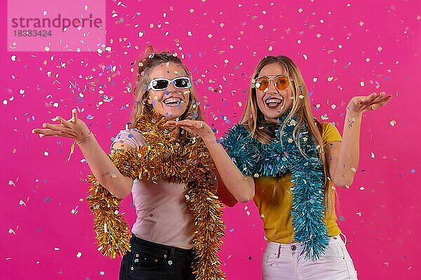 Zwei junge blonde kaukasische Frauen feiern in einem Nachtclub und haben Spaß beim Werfen von Konfetti  vor einem rosa Hintergrund