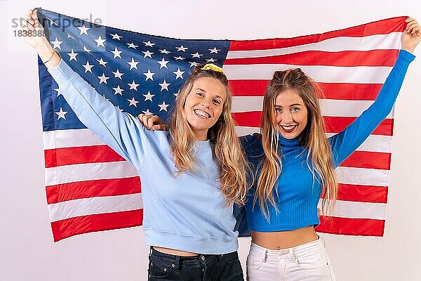 Zwei junge blonde kaukasische Frauen mit amerikanischer Flagge  lächelnd  während sie die Flagge halten  vor einem weißen Hintergrund