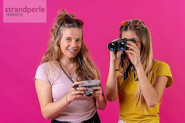 Zwei junge blonde kaukasische Frauen  Urlaub Porträt mit Kamera und Ferngläser  vor einem rosa Hintergrund