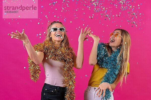 Zwei junge blonde kaukasische Frauen feiern und haben Spaß beim Konfettiwerfen  vor einem rosa Hintergrund