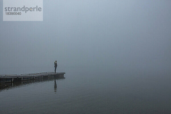 Blonde Frau steht an einem nebligen Morgen am Dock des Sees