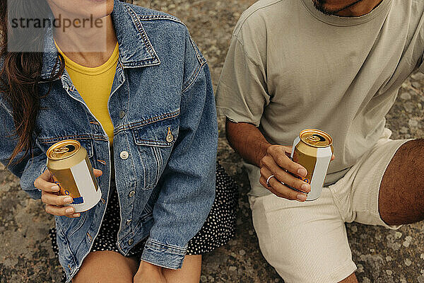 Mittelteil von männlichen und weiblichen Freunden  die Getränkedosen halten  während sie zusammen auf einem Felsen sitzen