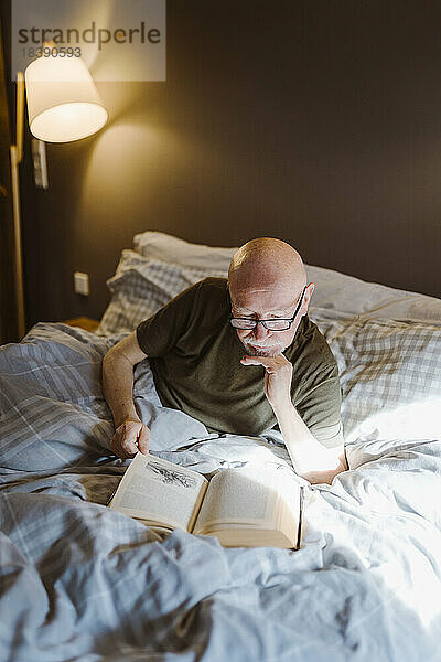 Älterer Mann mit Hand am Kinn liest ein Buch  während er zu Hause auf dem Bett liegt