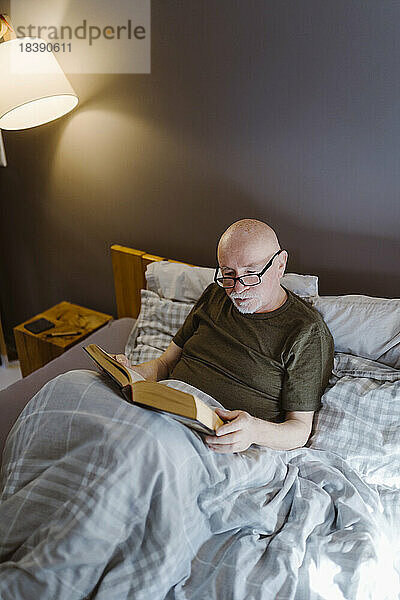 Älterer Mann mit Brille beim Lesen eines Buches auf dem Bett zu Hause