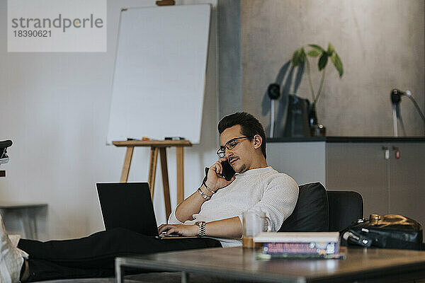 Junger männlicher Unternehmer  der mit seinem Smartphone telefoniert  während er an einem Laptop in einem kreativen Büro arbeitet