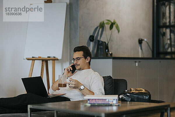 Geschäftsmann  der einen Kaffee trinkt  während er mit seinem Smartphone telefoniert und an seinem Laptop im Büro arbeitet