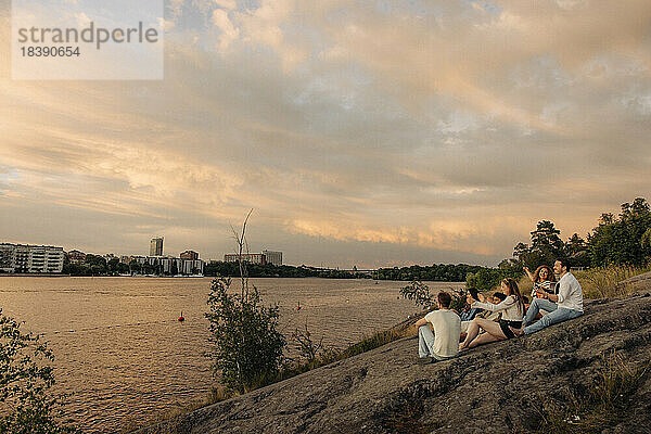 Multirassische männliche und weibliche Freunde genießen den Sonnenuntergang  während sie zusammen auf einem Felsen am Meer sitzen