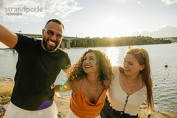 Glücklicher Mann genießt Picknick mit weiblichen Freunden gegen Meer bei Sonnenuntergang