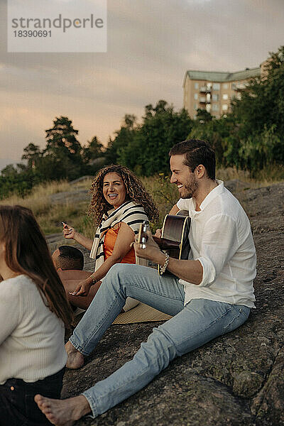 Lächelnder junger Mann spielt Gitarre  während er mit Freunden beim Picknick auf einem Felsen sitzt