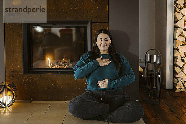 Frau mit geschlossenen Augen bei einer Atemübung im Schneidersitz am Kamin zu Hause