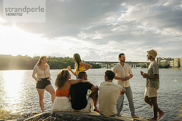 Multirassische männliche und weibliche Freunde unterhalten sich beim Picknick am Meer bei Sonnenuntergang