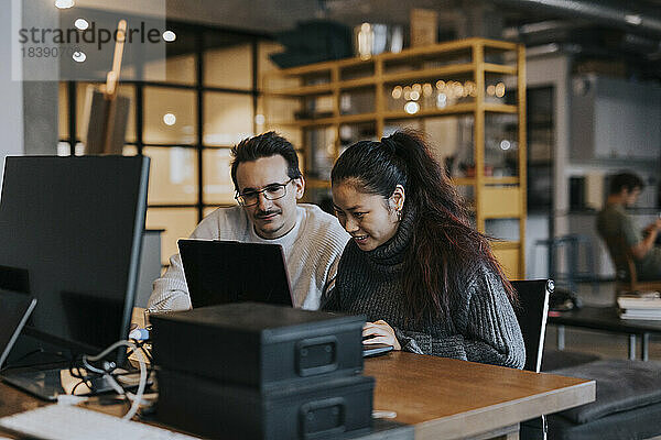 Lächelnde Geschäftsfrau arbeitet mit einem männlichen Kollegen an einem Laptop in einem kreativen Büro