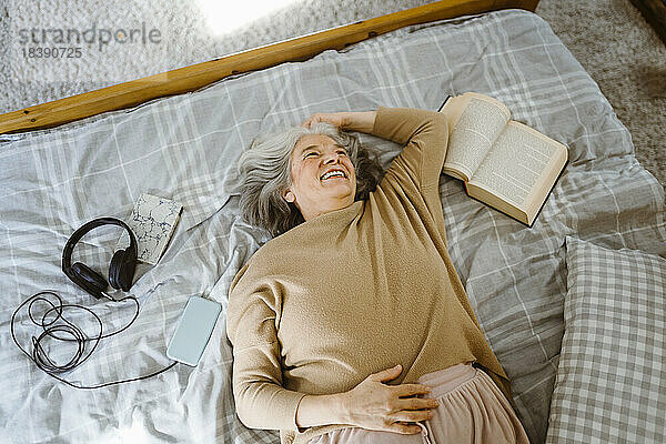 Glückliche ältere Frau mit Buch und Kopfhörer auf dem Bett liegend