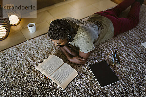 Hoher Blickwinkel eines Mannes  der Musik hört und ein Buch liest  während er zu Hause auf dem Teppich liegt
