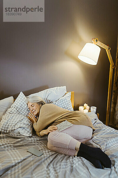 Ältere Frau schläft auf dem Bett in der Nähe einer beleuchteten Lampe zu Hause