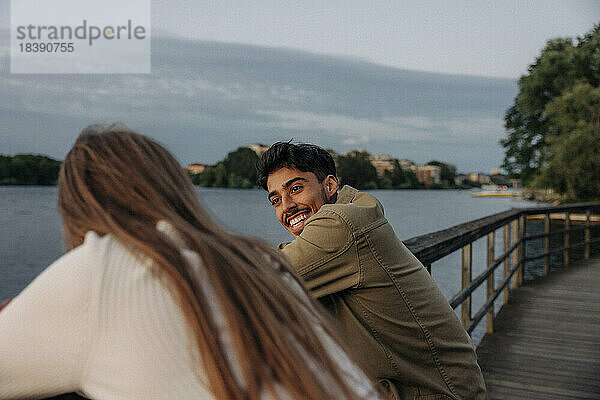 Seitenansicht eines glücklichen jungen Mannes  der sich mit einer Freundin unterhält  während er sich am Geländer eines Piers anlehnt