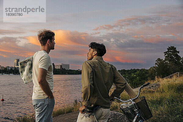 Seitenansicht eines jungen Mannes im Gespräch mit einem männlichen Freund mit Fahrrad bei Sonnenuntergang