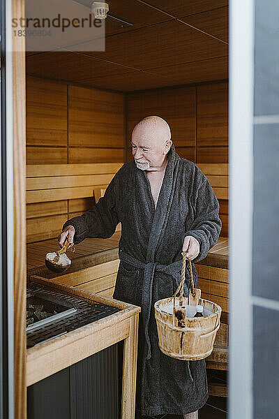 Ein älterer Mann legt einen heißen Stein in ein Heizgerät  während er in der Sauna steht