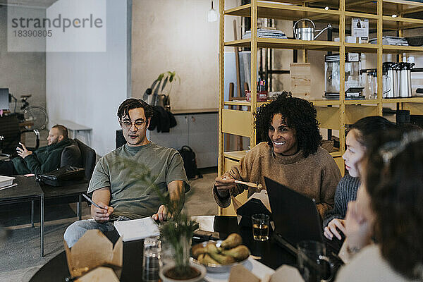 Lächelnde Geschäftskollegen  die beim Mittagessen am Tisch in einem kreativen Büro diskutieren