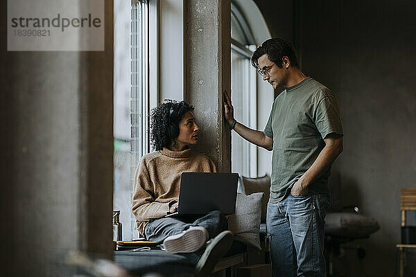 Eine Unternehmerin hält einen Laptop in der Hand  während sie mit einem männlichen Kollegen in einem Start-up-Unternehmen diskutiert