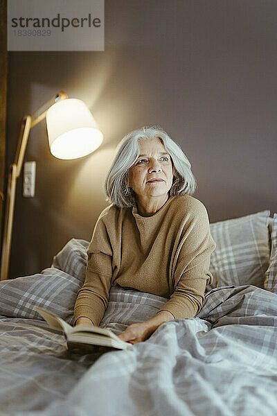 Nachdenkliche ältere Frau mit Buch  die zu Hause auf dem Bett sitzt