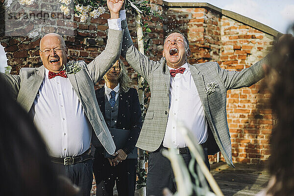 Glückliches schwules Paar  das sich an den Händen hält  während es die Hochzeitszeremonie eines Ministers an einem sonnigen Tag genießt