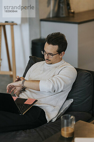 Junger Geschäftsmann  der die Zeit auf seiner Armbanduhr überprüft  während er mit seinem Laptop im Büro sitzt