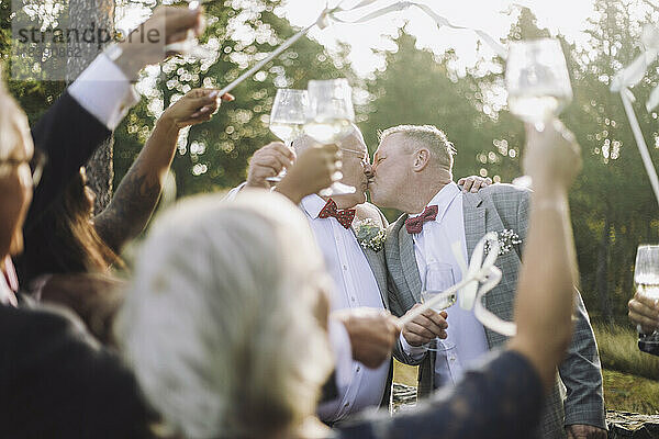 Neuvermähltes schwules Paar küsst sich vor den Gästen  die bei der Hochzeitszeremonie einen Toast ausbringen