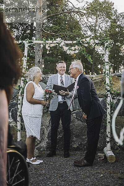 Älteres Paar tauscht das Eheversprechen aus  während der Hochzeitszeremonie steht der Pfarrer an einer Wand mit Blumenschmuck