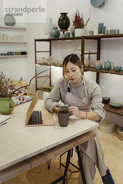 Junge Unternehmerin mit Laptop und Mobiltelefon in der Werkstatt sitzend