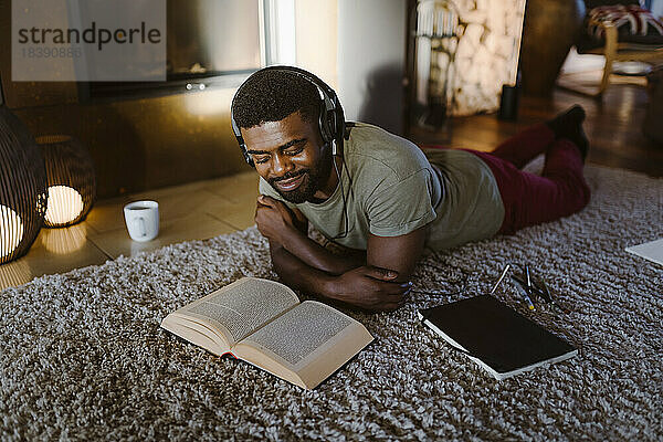 Lächelnder junger Mann  der Musik hört und ein Buch liest  während er zu Hause auf dem Teppich liegt