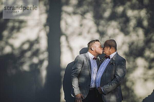 Zärtlicher schwuler Mann küsst glatzköpfigen Partner auf den Mund an der Wand während der Hochzeit an einem sonnigen Tag