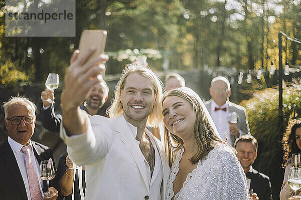 Glücklicher Bräutigam  der ein Selfie mit der Braut und den Gästen durch ein Smartphone bei der Hochzeitszeremonie macht