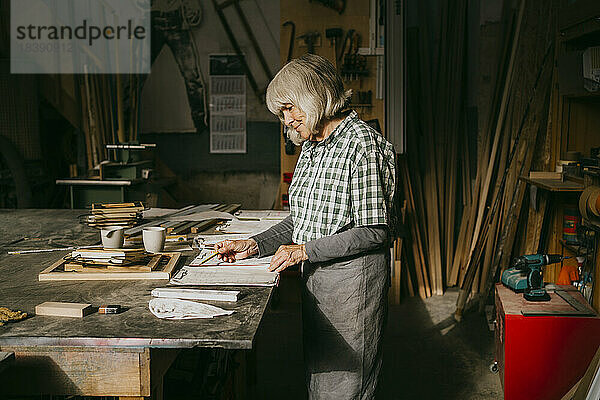 Seitenansicht einer älteren Schreinerin  die in der Werkstatt stehend ein Dokument liest