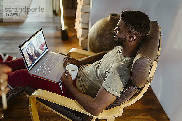 Lächelnder Mann führt Videogespräch mit Frau über Laptop  während er zu Hause auf einem Stuhl sitzt