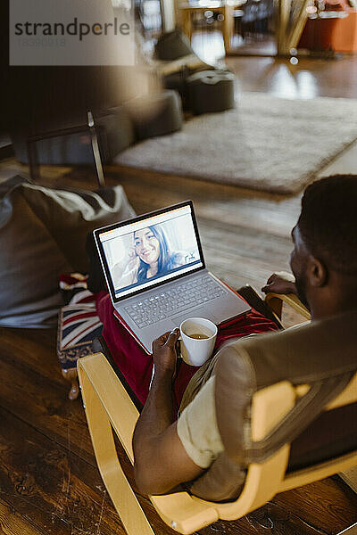 Mann führt Videogespräch mit Frau über Laptop  während er zu Hause auf einem Stuhl sitzt