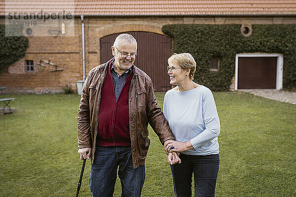 Lächelnde ältere Frau hält die Hand eines Mannes  der mit einem Stock auf einer Wiese im Hinterhof geht