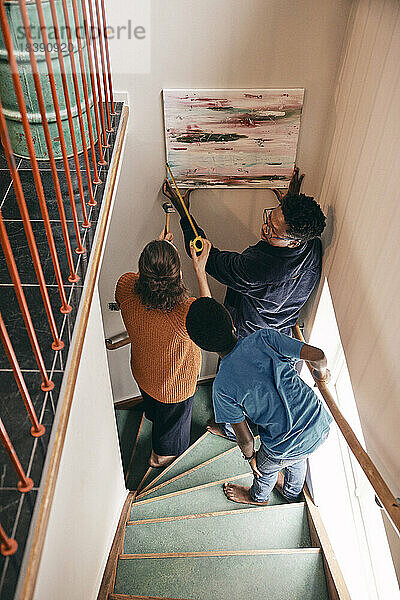 Blick von oben auf die Familie  die sich gegenseitig beim Aufhängen von Gemälden an der Wand zu Hause unterstützt