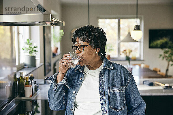 Ältere Frau in Jeansjacke trinkt Wasser in der Küche zu Hause