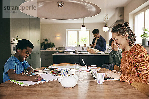 Glückliche Mutter mit Laptop und Sohn  der in einem Buch malt  während er zu Hause am Esstisch sitzt