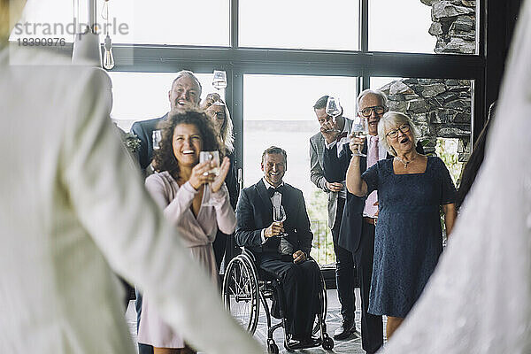 Glückliche multirassische Gäste mit Weingläsern  die dem frisch vermählten Paar bei der Hochzeitszeremonie zujubeln