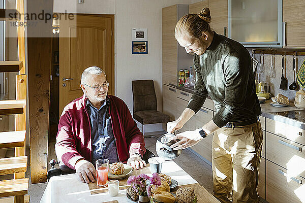 Junger männlicher Hausmeister gießt Kaffee für einen älteren Mann im Ruhestand mit Behinderung beim Frühstück zu Hause ein