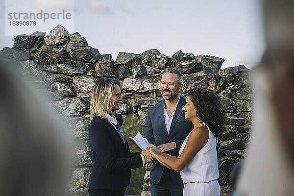 Glückliches lesbisches Paar  das sich an den Händen hält  während der Pfarrer sie während der Hochzeitszeremonie an der Steinmauer betrachtet