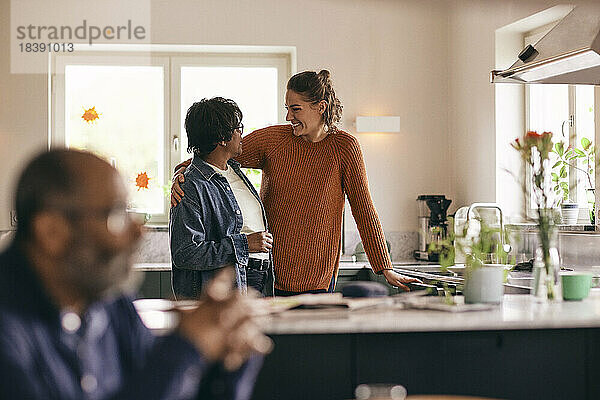 Glückliche Schwiegertochter  die den Arm um eine ältere Frau legt  die zu Hause in der Küche steht
