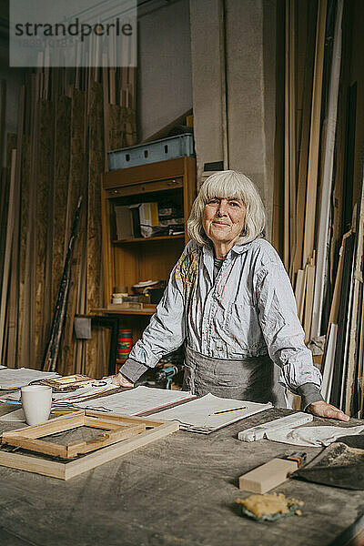 Lächelnder älterer weiblicher Schreiner  der sich in der Nähe der Werkbank in einer Werkstatt anlehnt