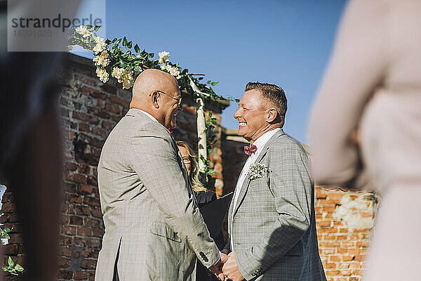 Glückliches frisch verheiratetes homosexuelles Paar  das sich bei der Hochzeit an den Händen hält  während eines sonnigen Tages