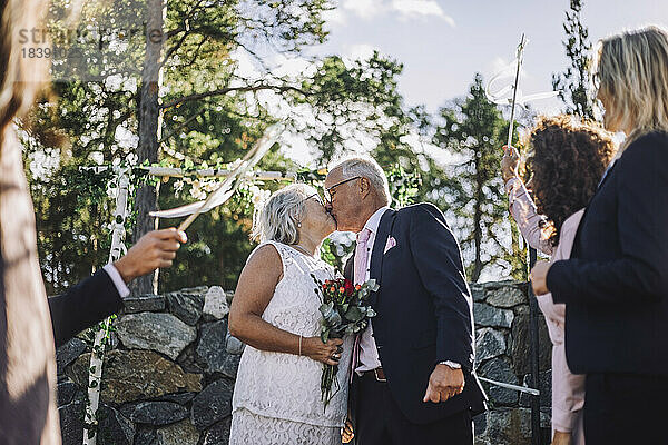 Ältere Braut mit Blumenstrauß küsst Bräutigam auf den Mund inmitten der Gäste während der Hochzeitsfeier